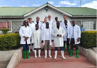Treffen mit Latia Agribusiness Solutions (LAS) Kenya 12 Personen mit weißen Kitteln und grünen sowie weißen Gummistiefeln stehen im Freien auf einer Treppe vor einem niedrigen Gebäude mit grünem Dach.