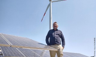 Peter Fischer steht neben einer Photovoltaikanlage. Hinter ihm ist ein Windrad zu sehen.